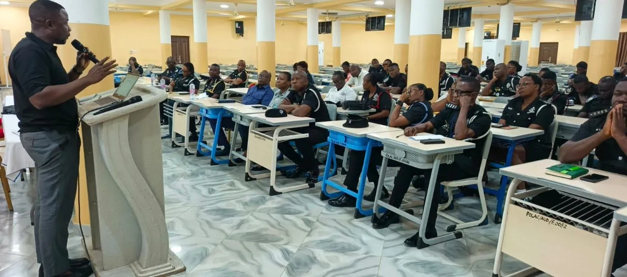 Uniformed Ghana Police Service officers seated at desks in a lecture hall, listening to a speaker presenting from a lectern.