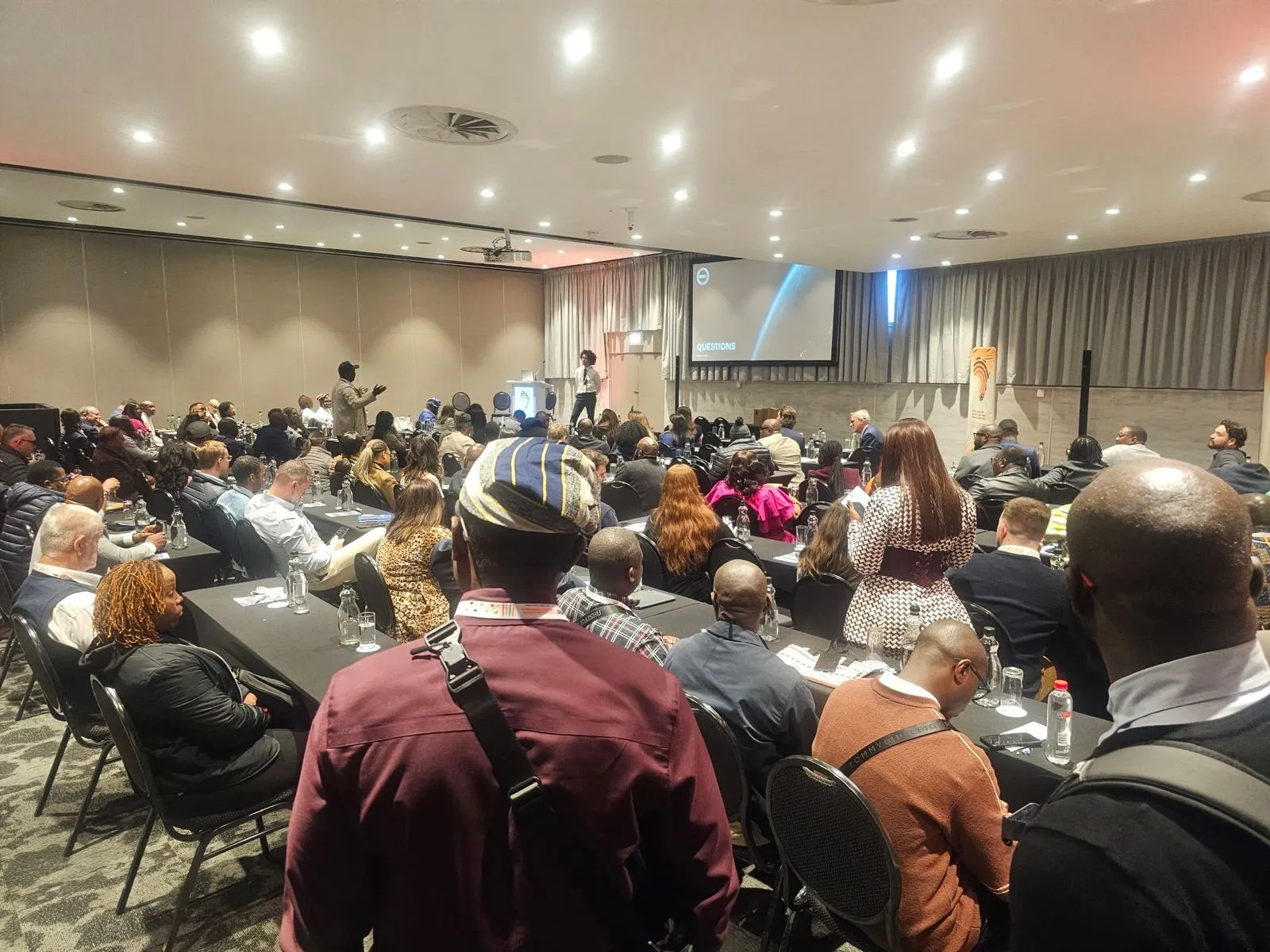A full conference room during a question, with a delegate standing to speak and the presenters listening from the front.